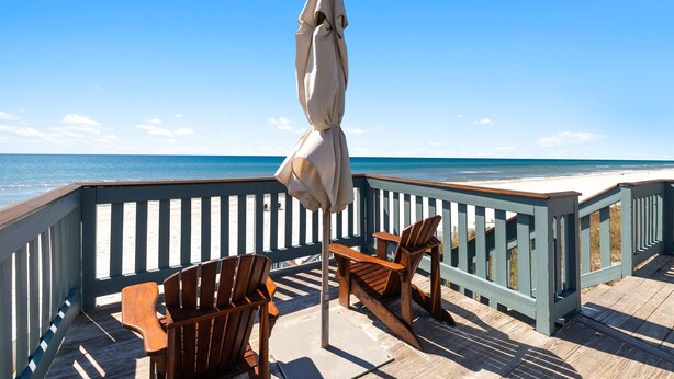A wooden deck with a railing, two wooden chairs, and a beach view.