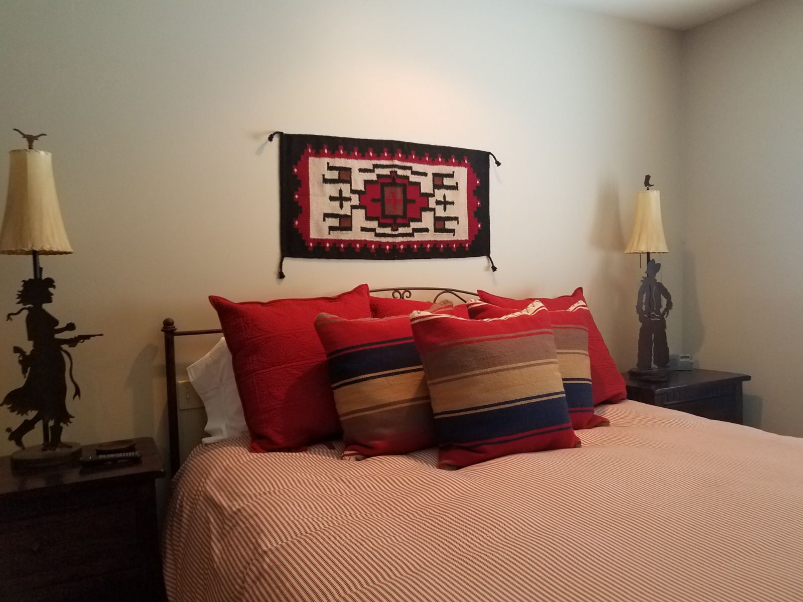 A neatly made bed with red and striped pillows, a patterned wall hanging, and two wall-mounted lamps on either side.