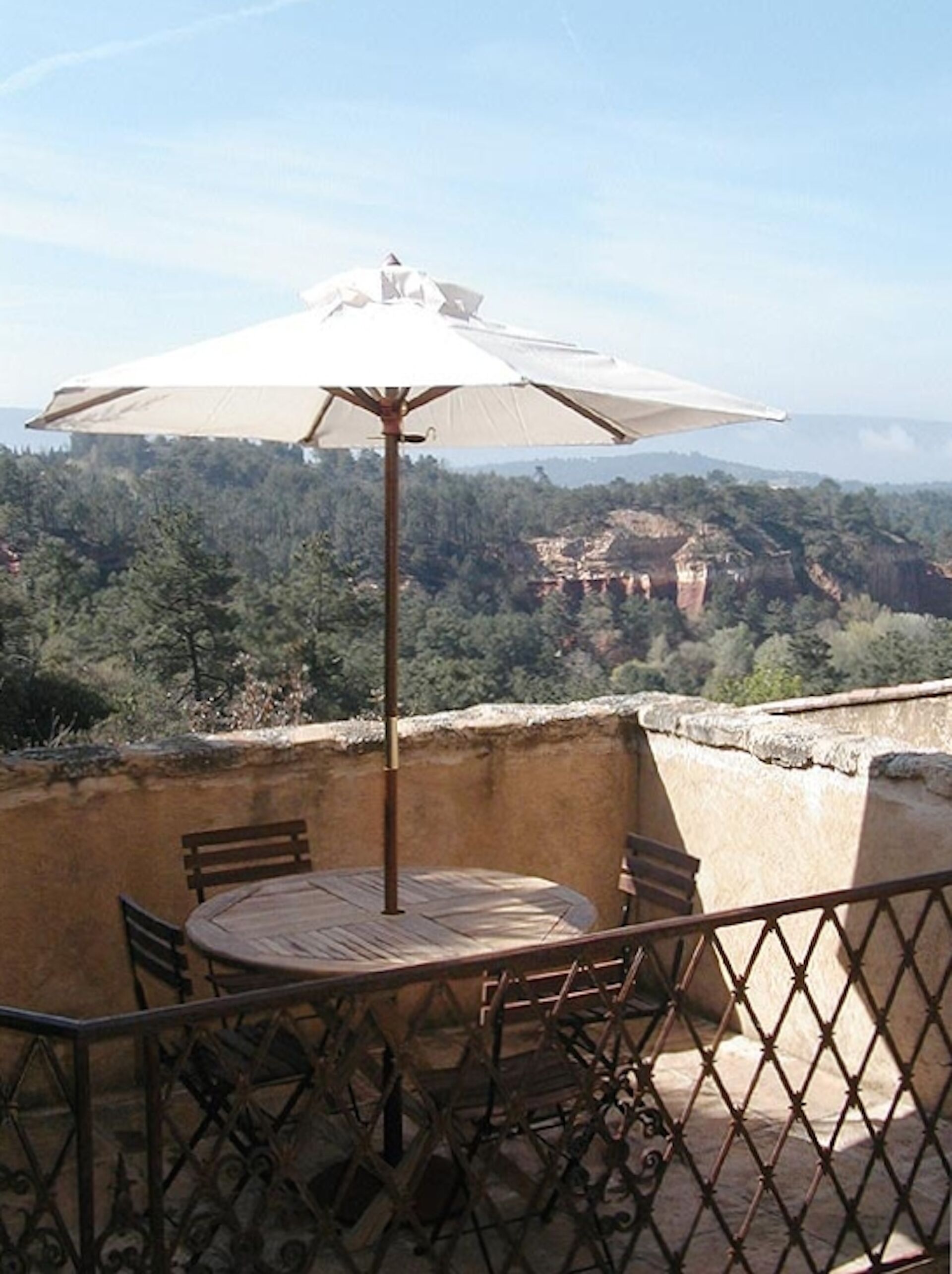 A balcony with a round table and chairs, an umbrella, and a scenic view of a forested area and rocky cliffs.