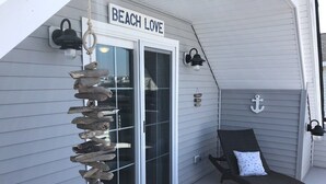 Un porche cubierto con un letrero que dice "BEACH LOVE", una decoración de leña, una silla de descanso con cojín y un adorno de ancla.