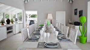 Un comedor con una mesa larga dispuesta para una comida, sillas blancas y una planta verde en maceta.