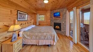 A cozy wooden cabin room with a bed, a fireplace, a TV, and a window with curtains. - Free Tickets | Secluded Spacious Cabin w/Mountain View, Hot Tub & Game Room (Sevierville)