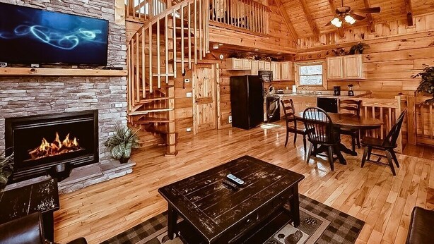 A spacious log cabin interior with a fireplace, a TV, a dining area, and a staircase leading to an upper level. - Free Tickets | Secluded Spacious Cabin w/Mountain View, Hot Tub & Game Room (Sevierville)