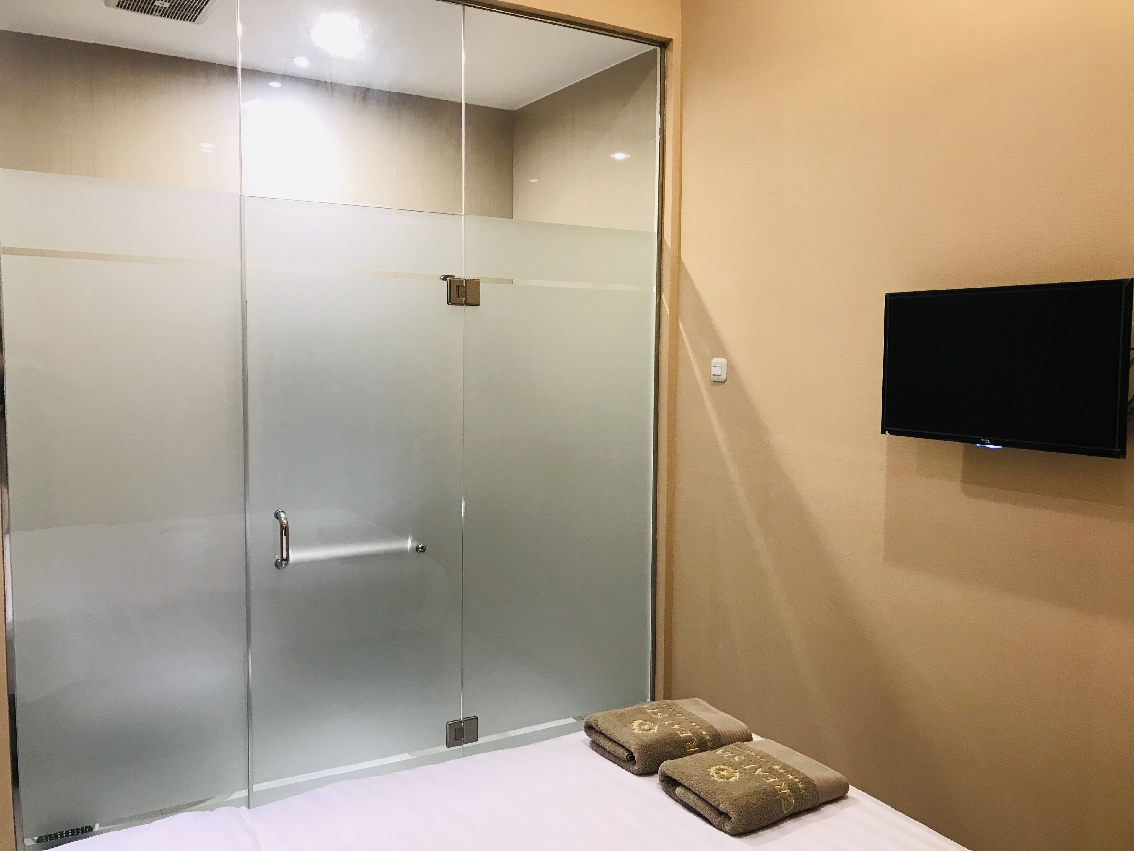 A hotel room with a glass-enclosed shower, a flat-screen TV mounted on the wall, and two towels on the bed.