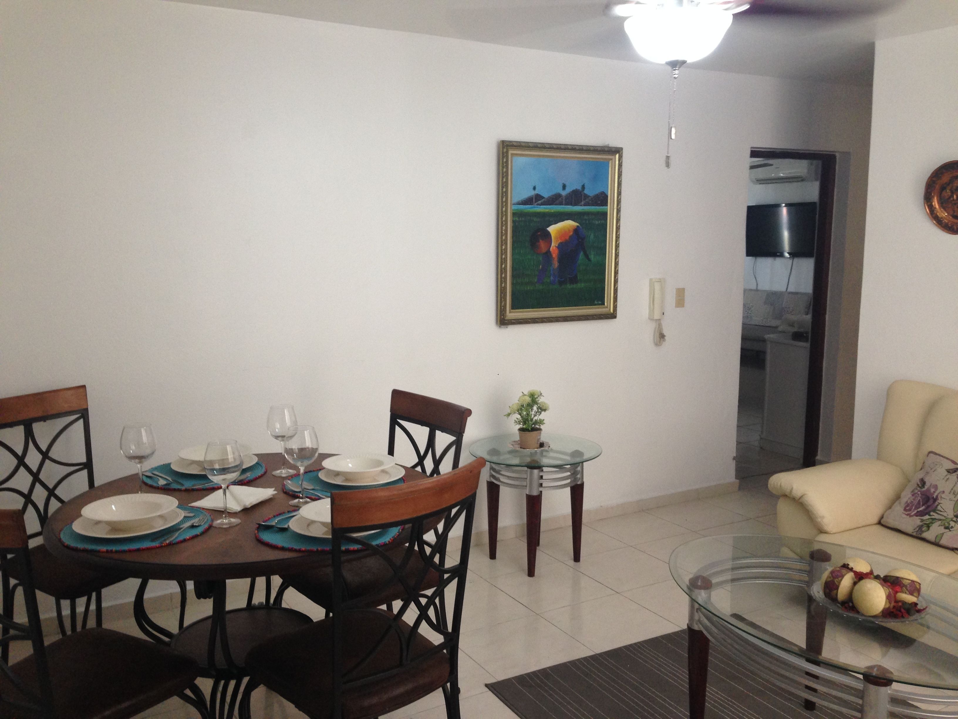A dining area with a round table set for four, a sofa, a small glass table with a bowl of fruit, and a painting on the wall.