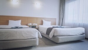 Habitación de hotel con dos camas, cabecero de madera, mesita de noche con lámpara y ventana grande con cortinas traslúcidas.