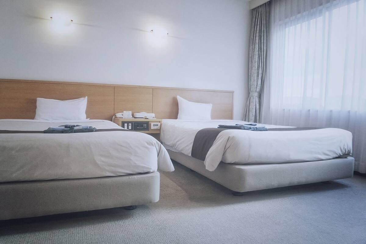 A hotel room with two beds, a wooden headboard, a bedside table with a lamp, and a large window with sheer curtains.