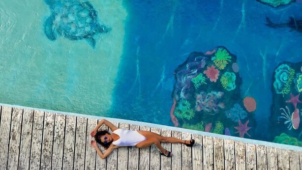 Una mujer recostada en una cubierta de madera junto a una piscina con agua cristalina y una tortuga nadando cerca.