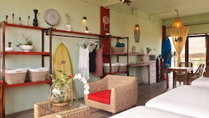 Una habitación con una cama, una silla de mimbre con un cojín rojo, una mesa con superficie de vidrio, estanterías con plantas y una tabla de surf, y un comedor con mesa y sillas.