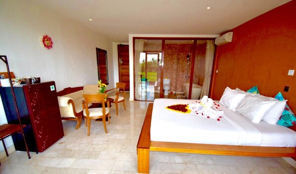 A hotel room with a large bed, a desk with a chair, a small table with a vase of flowers, and a sliding glass door leading to a balcony.
