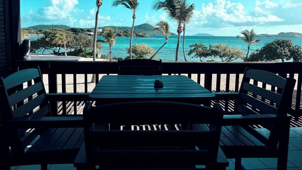 Une terrasse en bois avec une table et des chaises, donnant sur une plage bordée de palmiers et sous un ciel bleu azur.