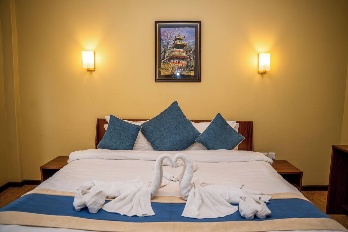 A hotel room with a bed, pillows, a framed painting, and wall sconces.