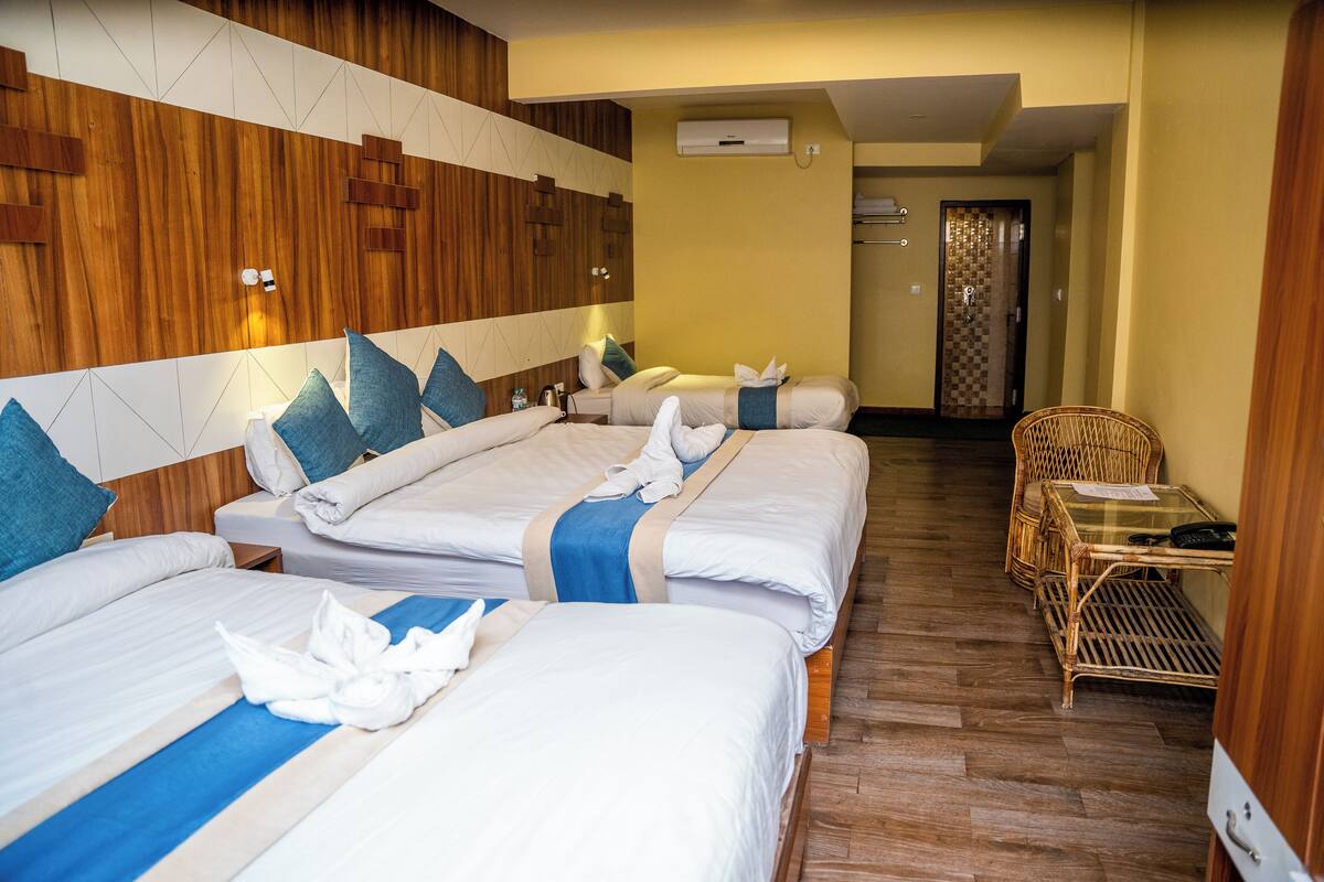 A hotel room with three beds, wooden paneling on the walls, a wicker chair, and a small table with a remote control.