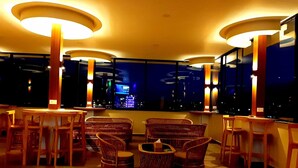 Ett modernt loungeområde med runda bord och fåtöljer i rotting, stora golvlampor och utsikt över stadsbilden på natten.