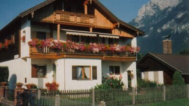 A two-story chalet with a balcony adorned with flowers, set against a mountainous backdrop. - Ferienwohnung für 4 Personen (75 qm) (Kiefersfelden)