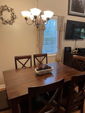 A dining area with a wooden table, four chairs, a chandelier, a basket of pinecones, a television, and a window with blinds. - Welcome to our country cabin! Sleeps 6. (Dahlonega)