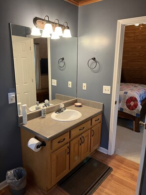 A bathroom with a large mirror, a sink, wooden cabinets, a towel rack, and a door leading to a bedroom. - Welcome to our country cabin! Sleeps 6. (Dahlonega)