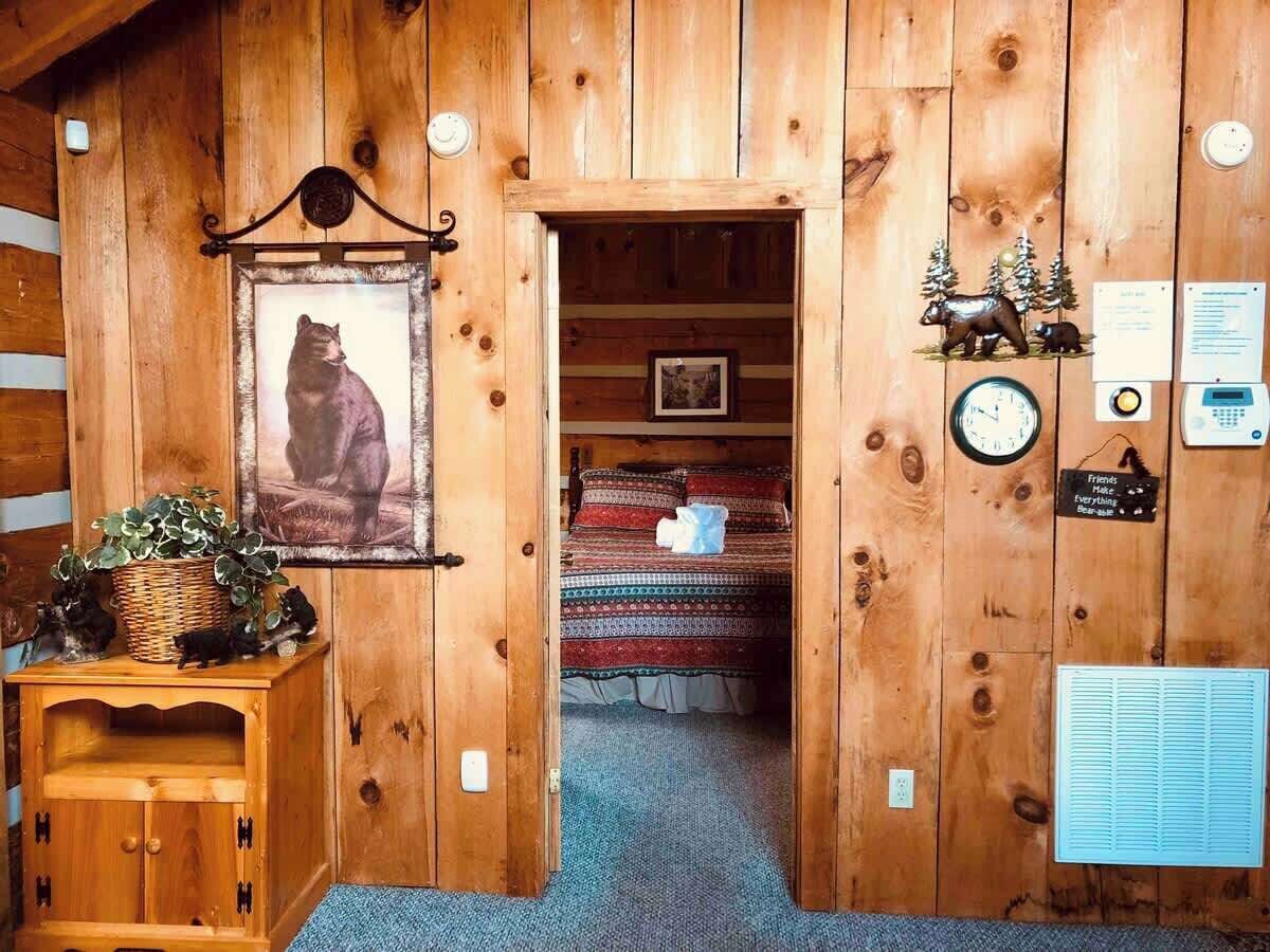 A wooden cabin interior with a bed, a framed picture of a bear, a basket with plants, and a small cabinet.