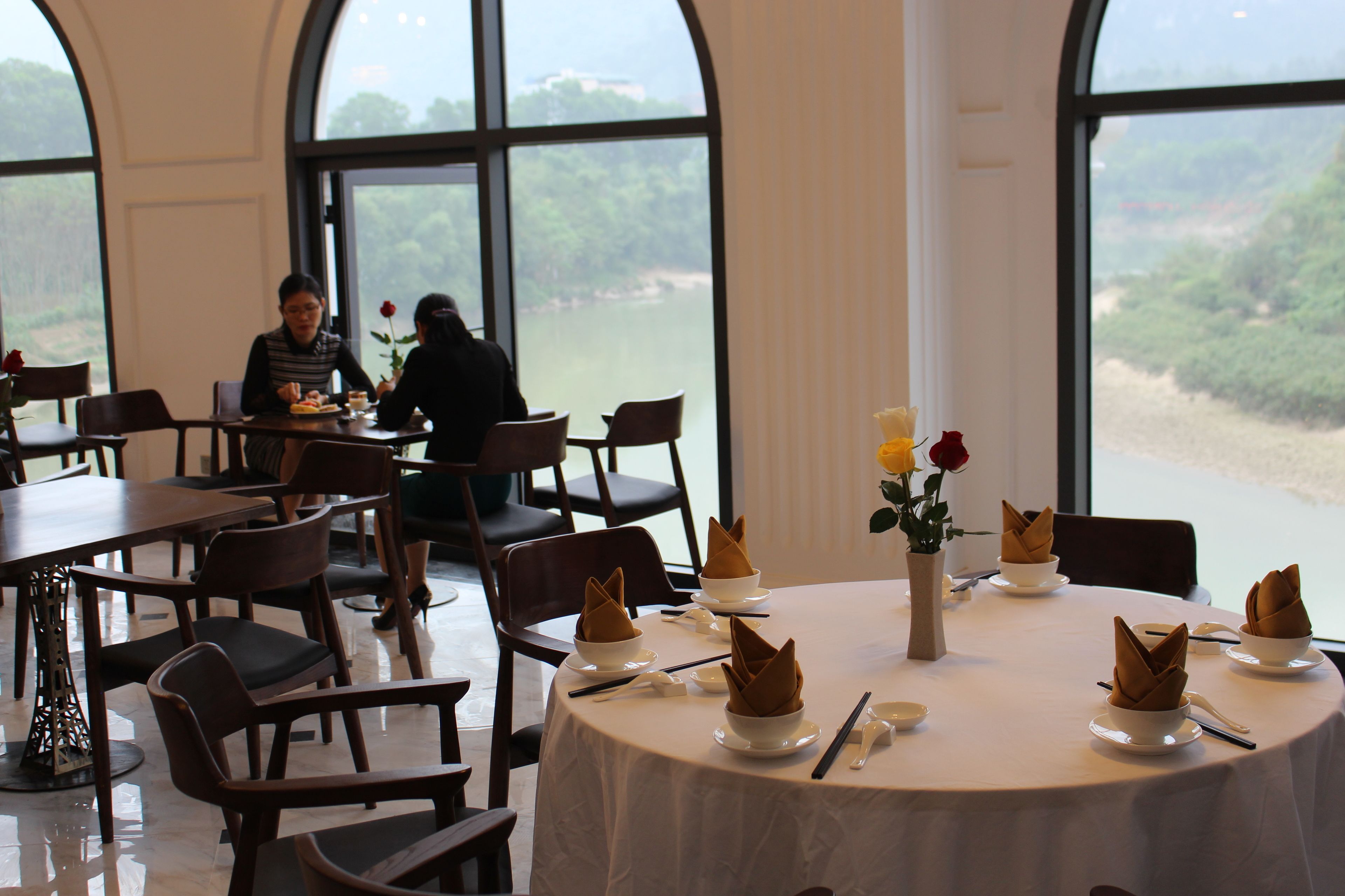 Un comedor con mesas dispuestas para dos, ventanas amplias que ofrecen vistas al exterior, y un jarrón con flores sobre la mesa.