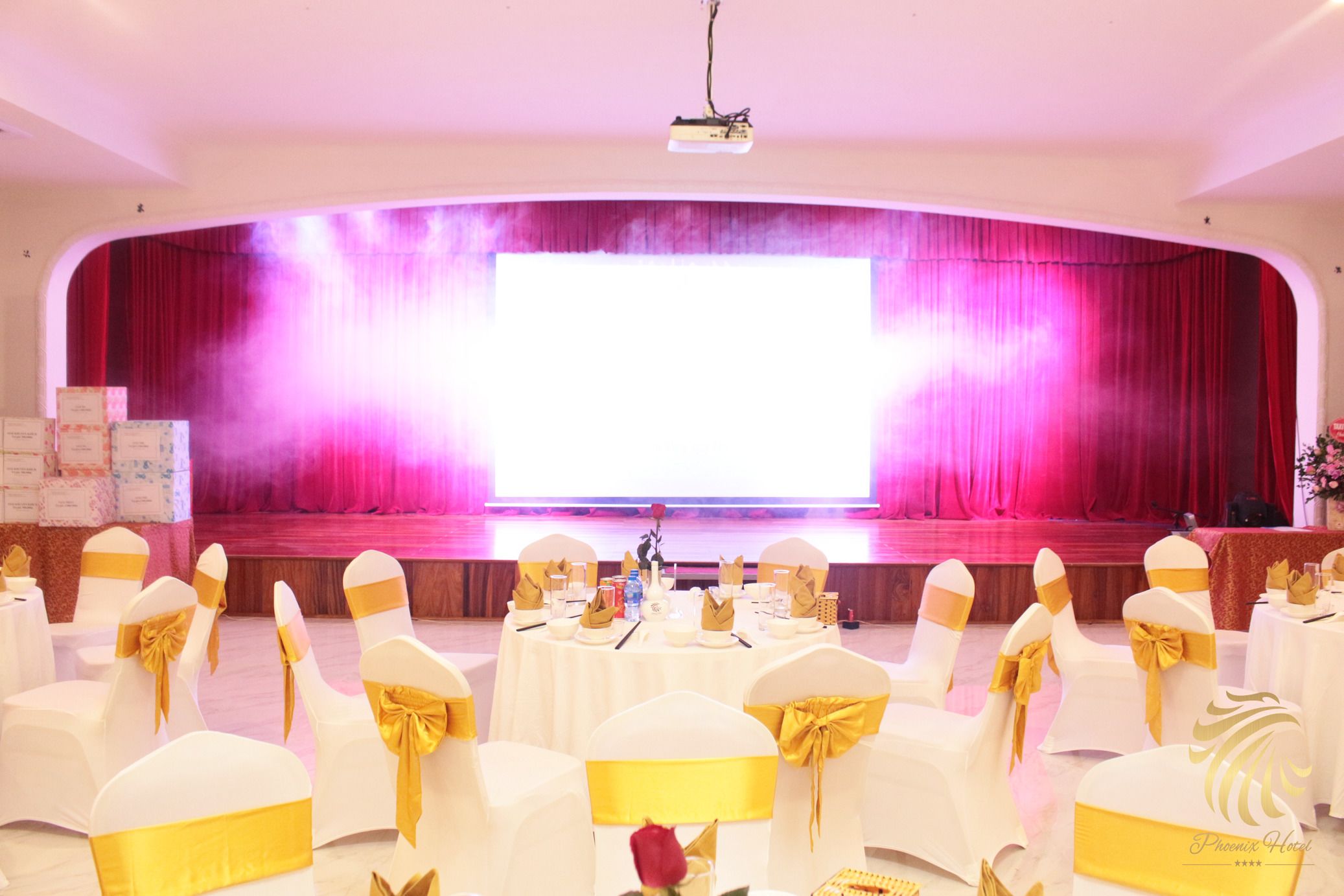 Un salón de conferencias con filas de sillas blancas con cintas amarillas, un escenario con cortina rosa y un proyector montado en el techo.