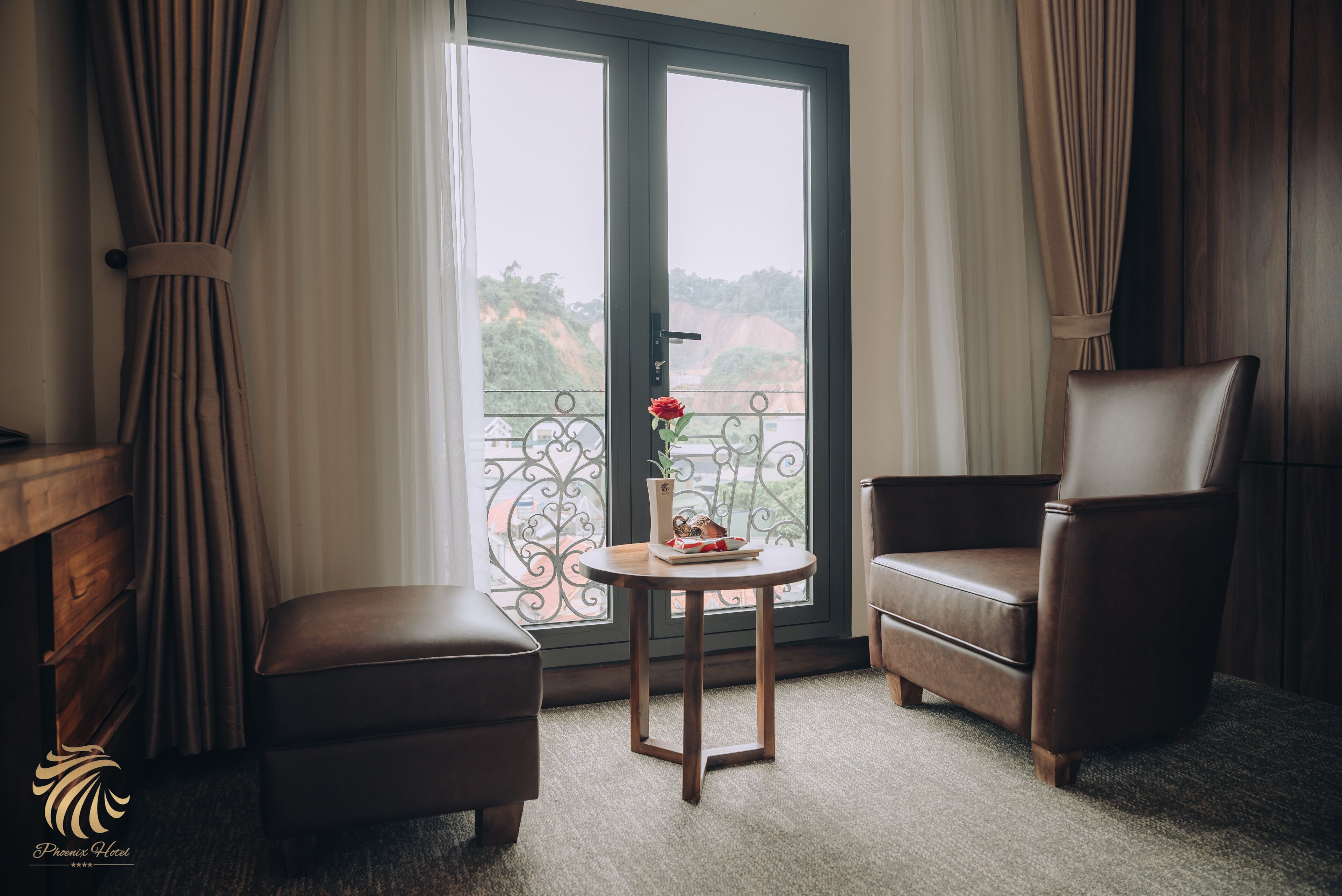 Une chambre d’hôtel avec un balcon, une table en bois ornée d’un vase de fleurs, deux fauteuils en cuir et une armoire en bois.