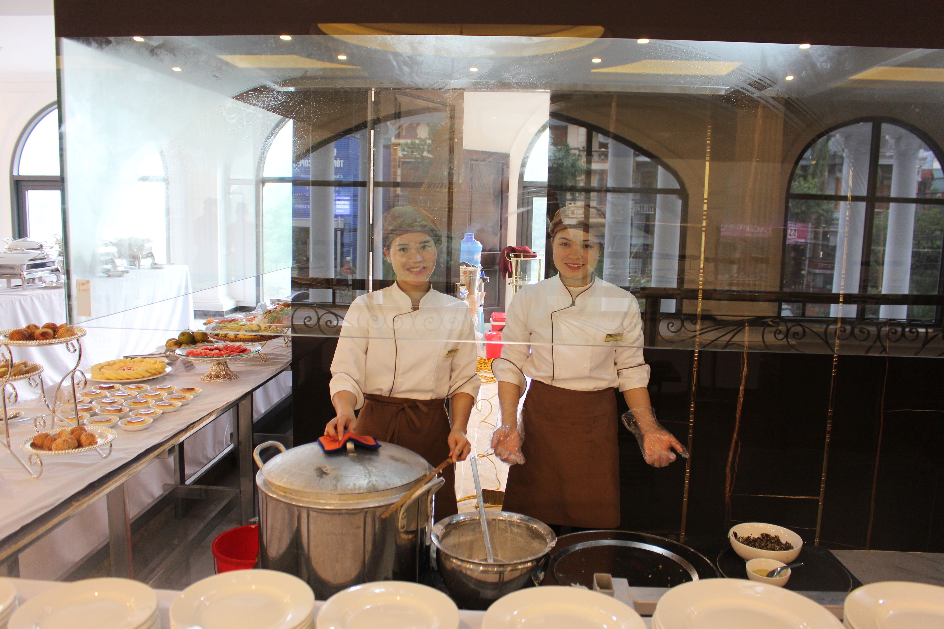 A kitchen with chefs preparing food, a buffet with various dishes, and large windows allowing natural light.