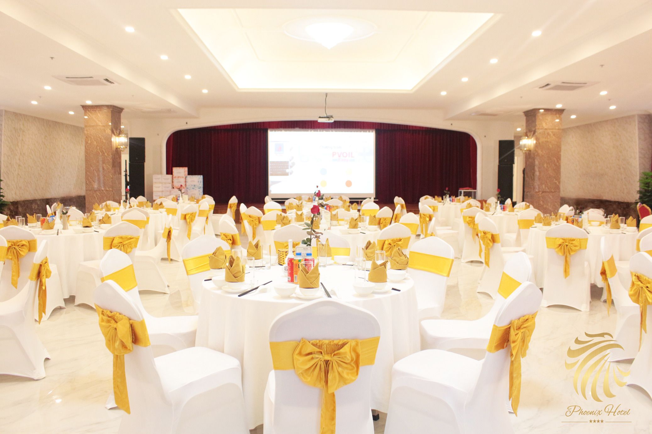 Un salón de banquetes con mesas redondas, sillas blancas con cintas amarillas y un escenario con pantalla de presentación.