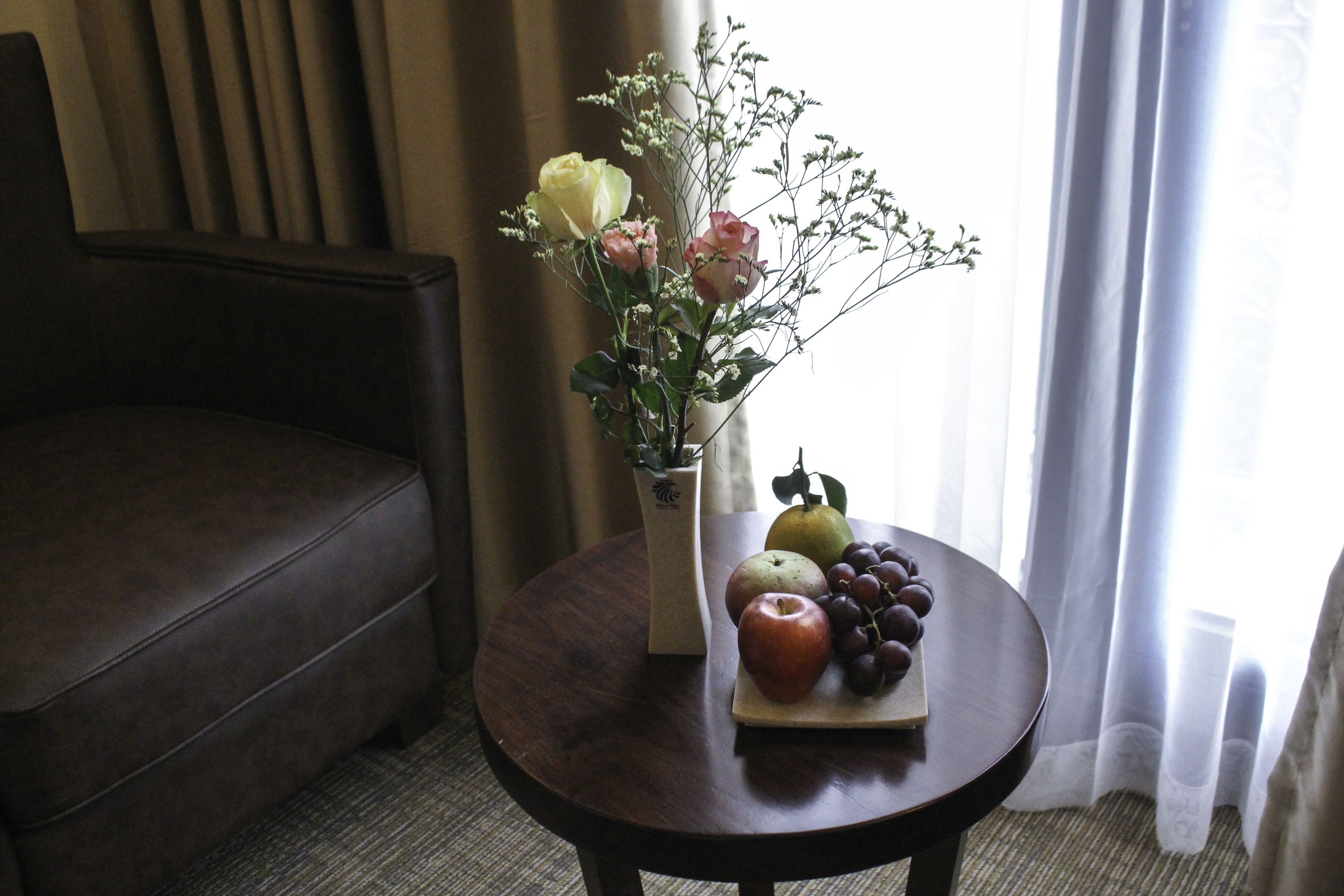 A round wooden table with a vase of flowers, a fruit bowl, and a vase with a plant.