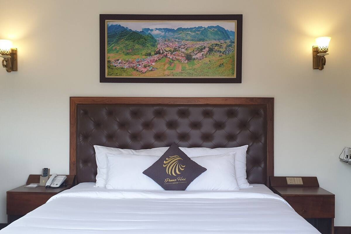 Une chambre d’hôtel avec un grand lit, une tête de lit en bois, deux tables de chevet avec des lampes, un tableau encadré au-dessus du lit et une fenêtre avec un fer à repasser.