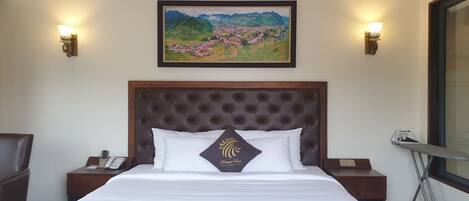 A hotel room with a large bed, a wooden headboard, two bedside tables with lamps, a framed painting above the bed, and a window with an ironing board.