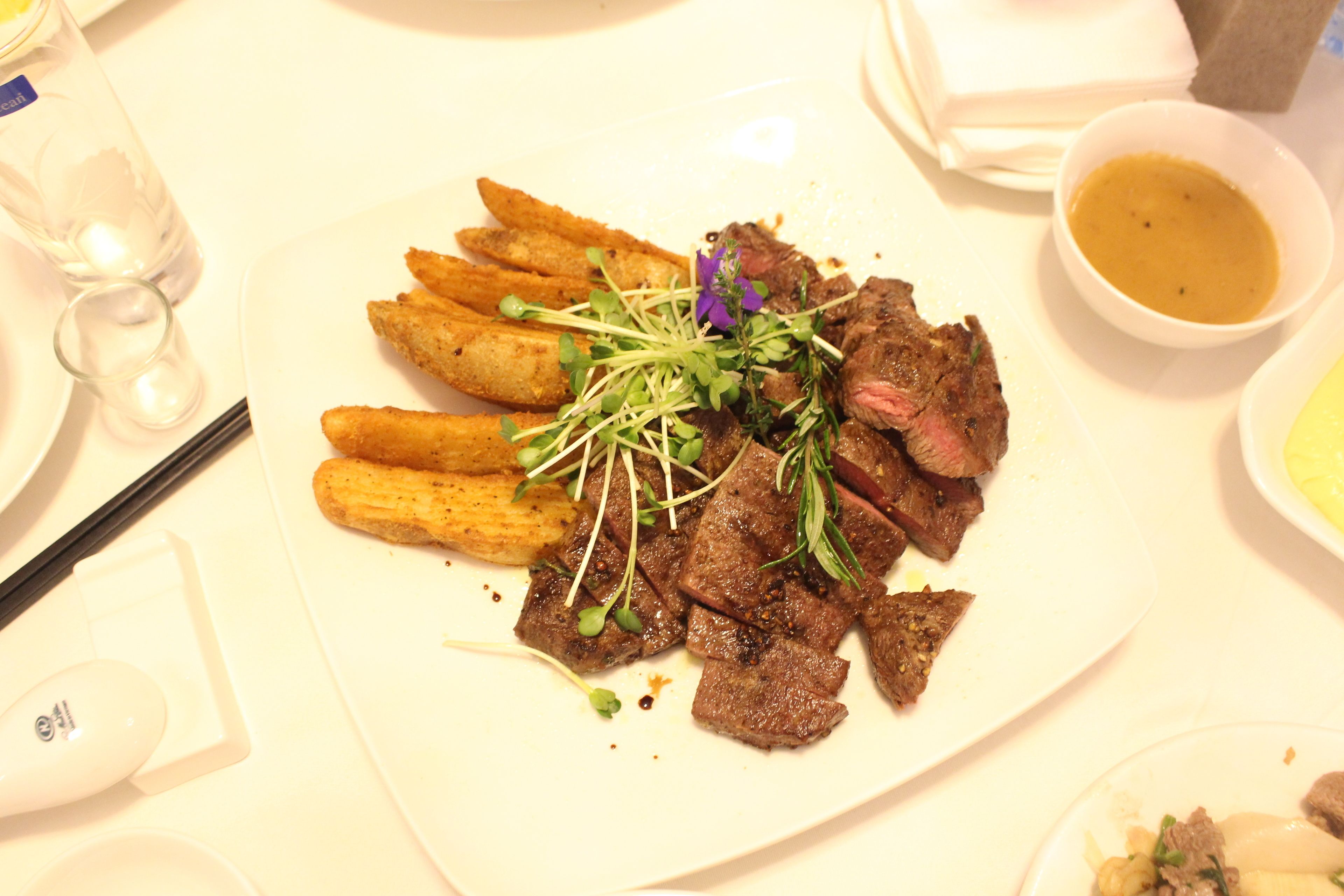 A plate of food with steak and fries, a small bowl of sauce, and a glass of water.
