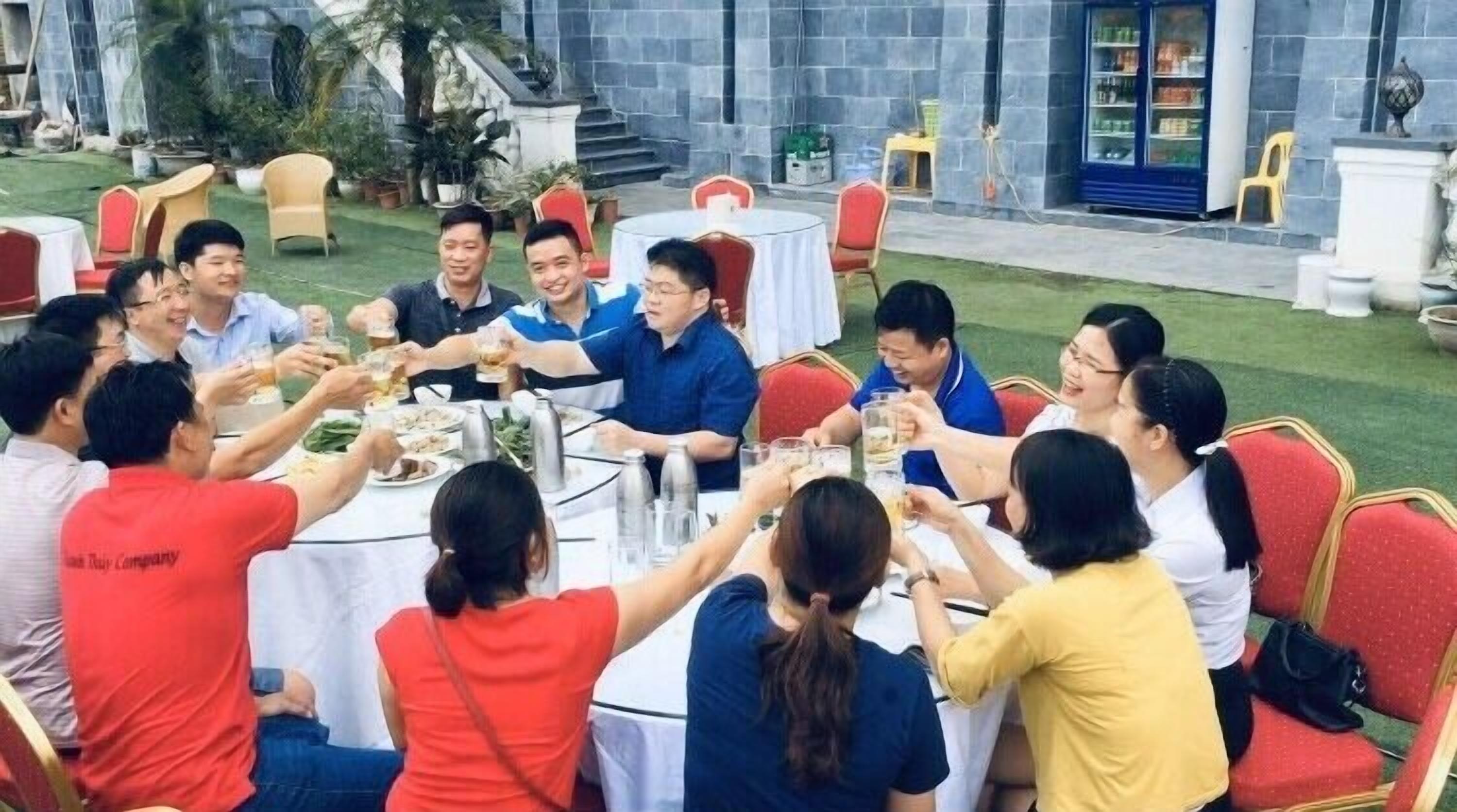 A group of people toasting around a table in an outdoor setting with a stone building in the background.
