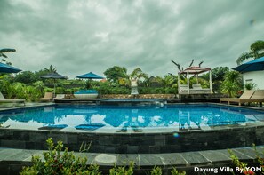 Una piscina con sillas de descanso y sombrillas, rodeada de exuberante vegetación y un cielo nublado.