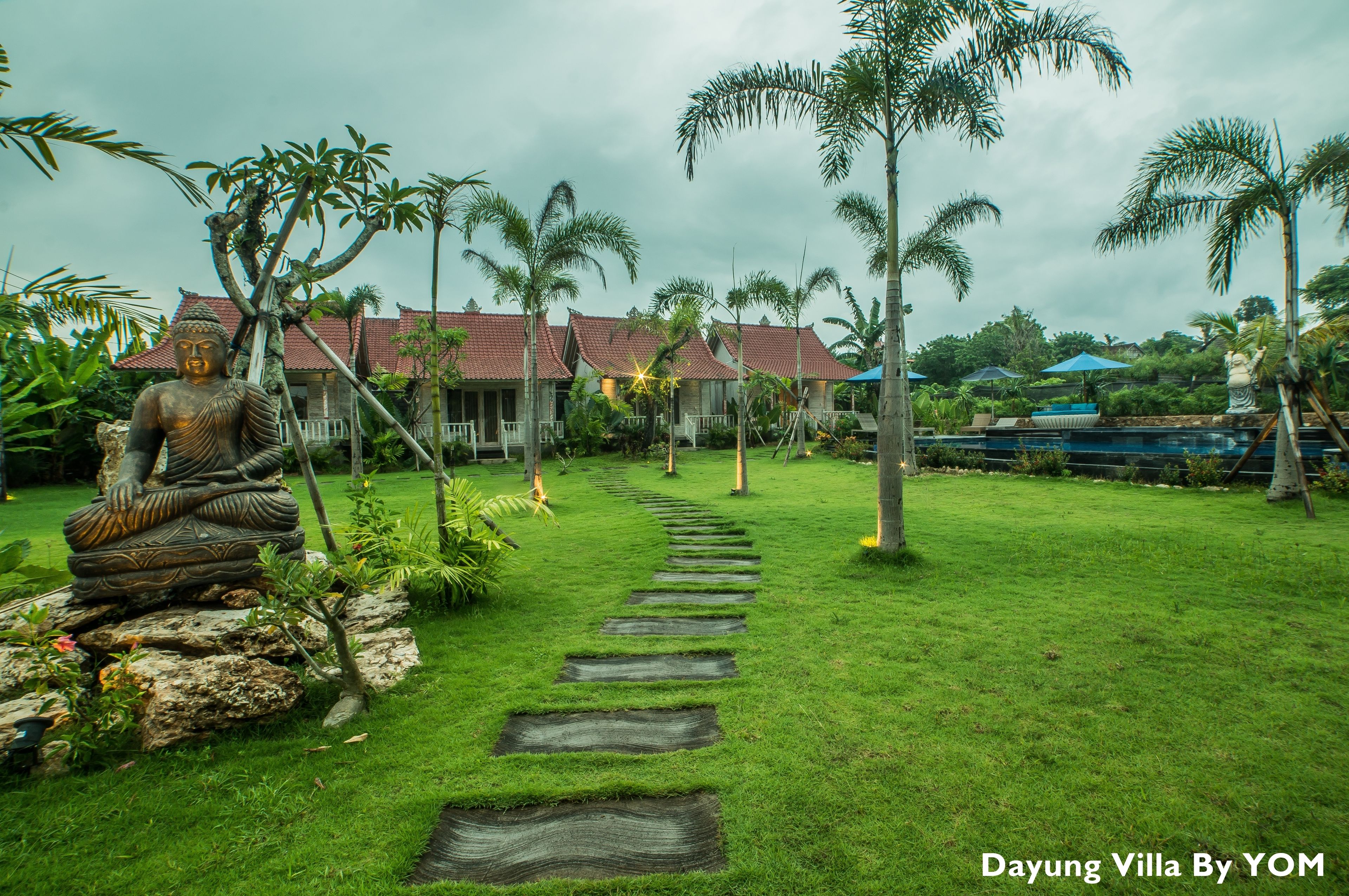 A Buddha statue in a garden with a pathway leading to a building with a red roof.
