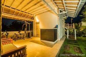 Un espacio exterior techado con equipo de ejercicio, una mesa de recepción y una escalera que lleva a un jardín.