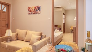 A hotel room with a beige sofa, a dining table set with plates, glasses, and a bottle. There is a bed in the background and a painting on the wall. - Chrissomallis Apartments (Skiathos)