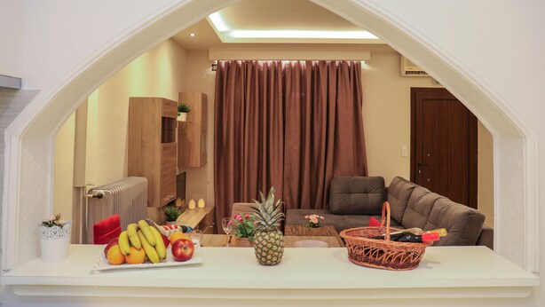 A living room with a white archway, a brown curtain, a brown sofa, a wooden cabinet, and a table with fruits and a bottle. - Chrissomallis Apartments (Skiathos)