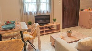 A cozy living room with a TV, a dining table with chairs, a sofa, and a kitchen area with cabinets and a fruit basket. - Chrissomallis Apartments (Skiathos)