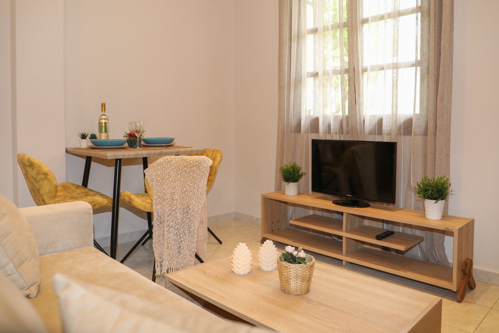 A living room with a sofa, a yellow chair, a wooden coffee table, a TV on a wooden stand, and a window with sheer curtains.