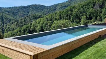 A rectangular swimming pool with a wooden deck, surrounded by a grassy area and a forested hillside.