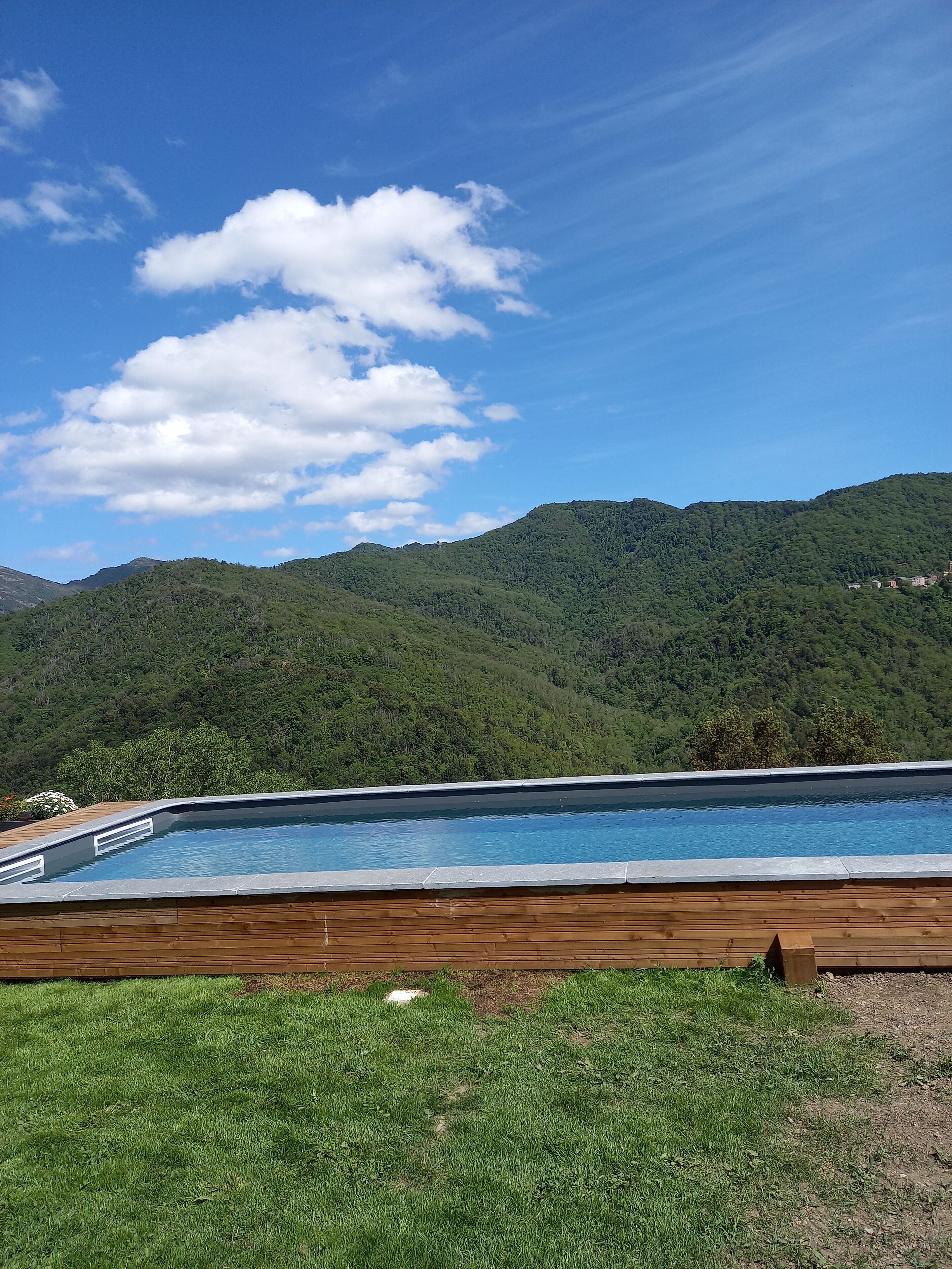 Una piscina con plataforma de madera, rodeada de colinas verdes y un cielo azul despejado.