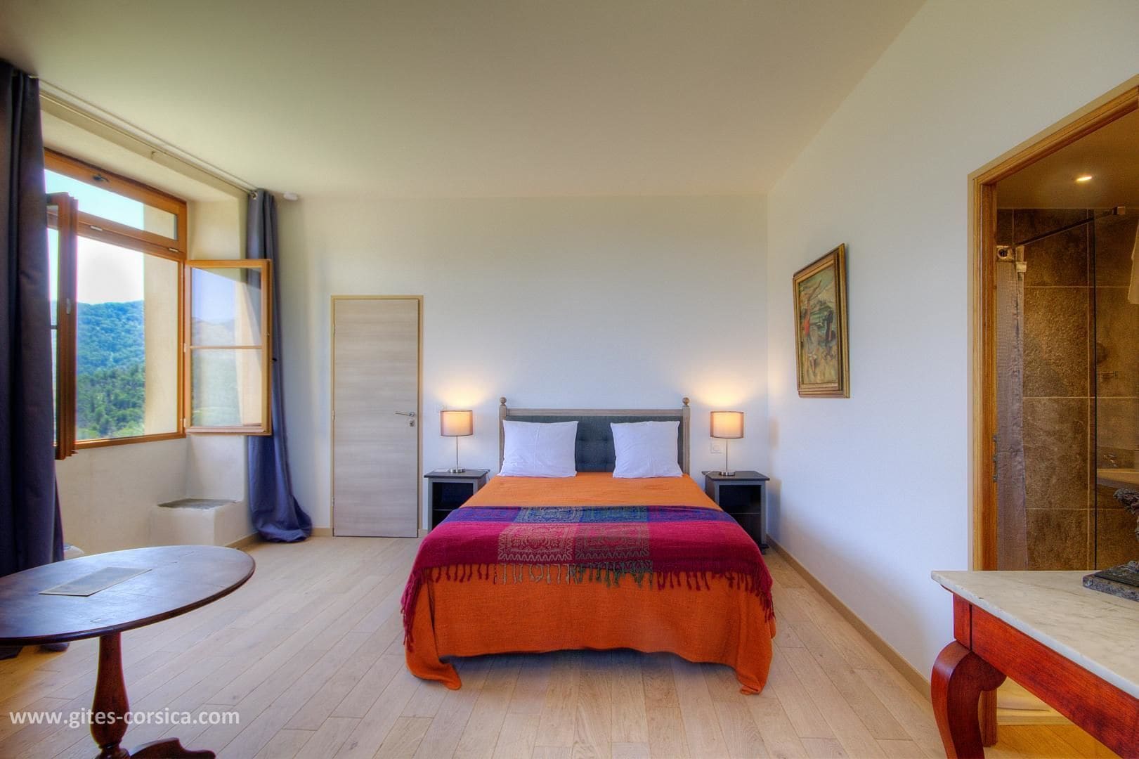 Ein Schlafzimmer mit Bett, Nachttischen, einem runden Tisch, einem Fenster mit Vorhängen und einer Tür.