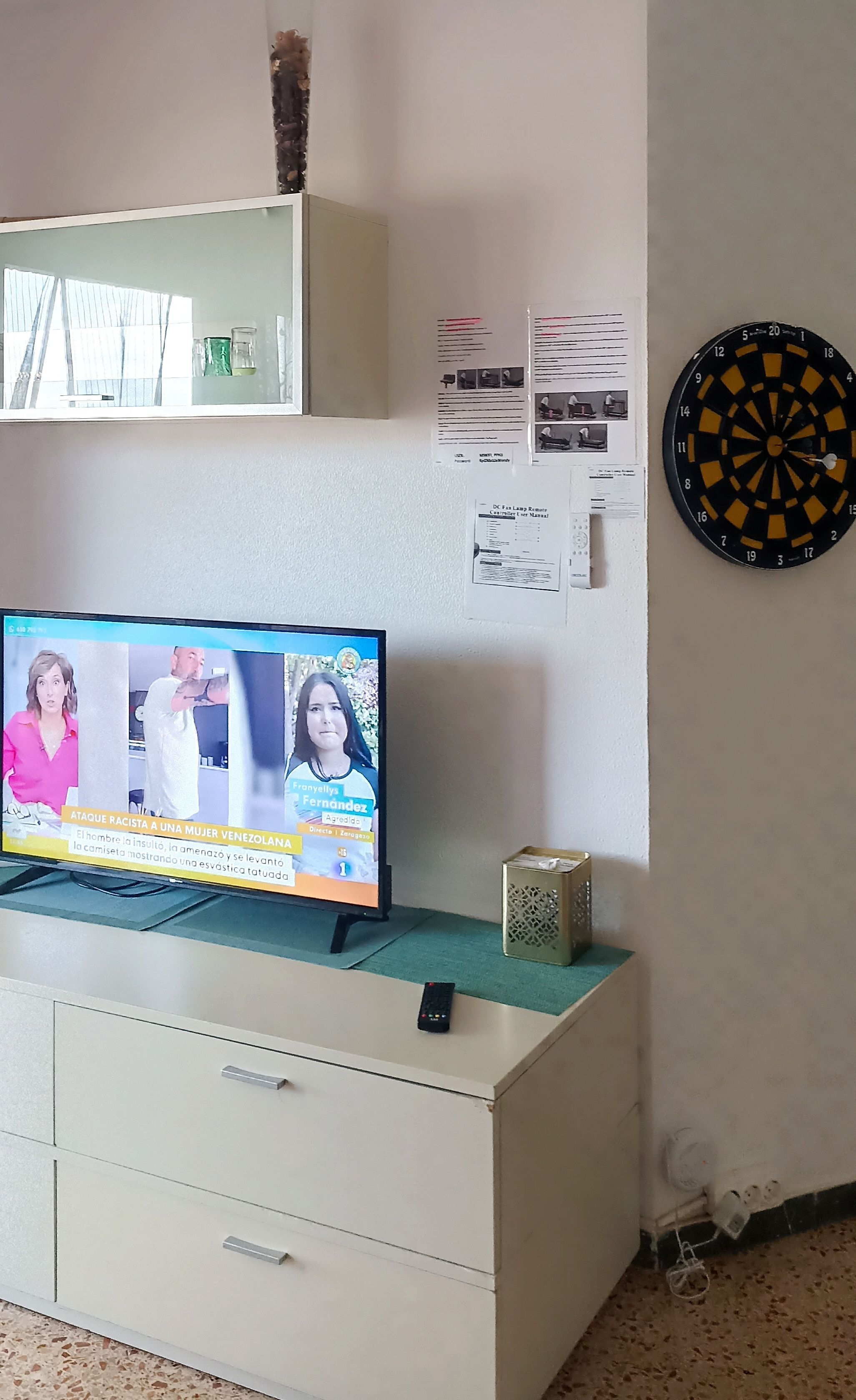 Uma televisão sobre um armário branco, um alvo de dardos e uma parede com papéis e um vaso.