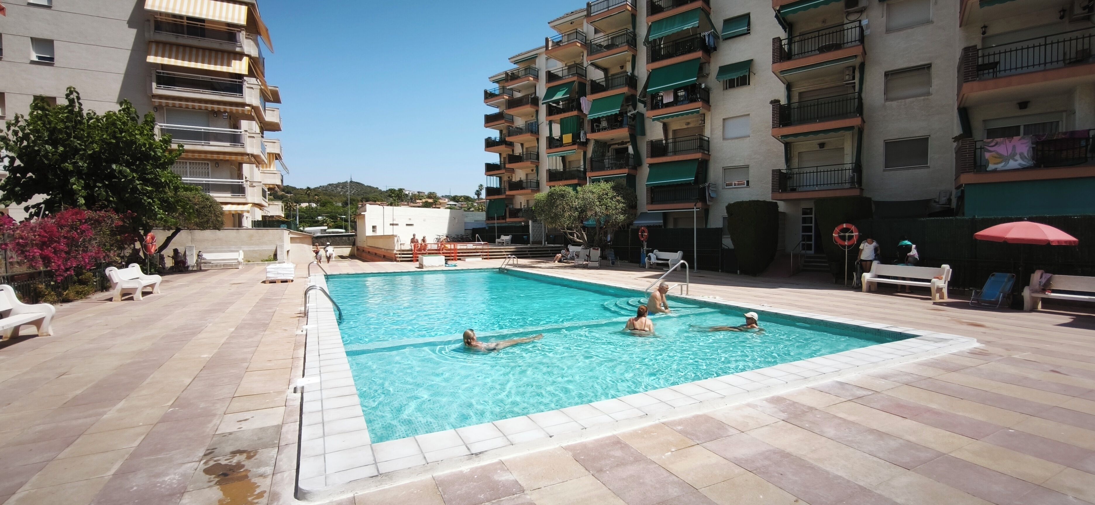 Una piscina rodeada de suelo de baldosas, con gente nadando y tomando el sol.