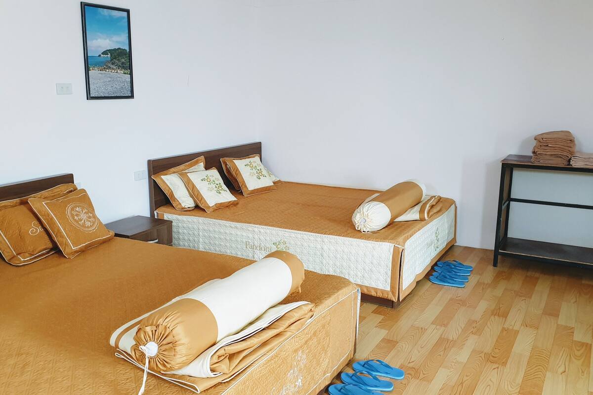 A hotel room with two beds, each with matching brown bedding and decorative pillows. There is a small table with folded towels and a pair of blue slippers on the floor.