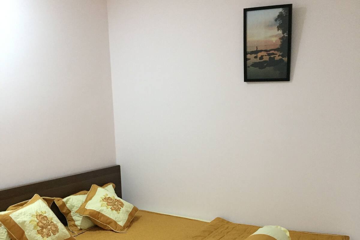 A neatly made bed with a brown bedspread and decorative pillows, a framed picture on the wall, and a plain white wall.