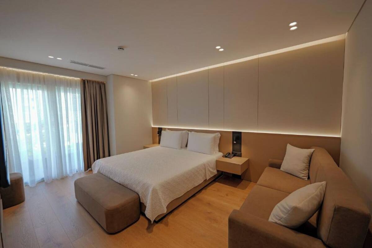 A modern hotel bedroom with a large bed, a sofa, and a bedside table.