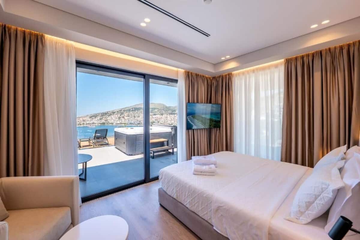A hotel room with a bed, a TV, a balcony with a hot tub, and a city view.
