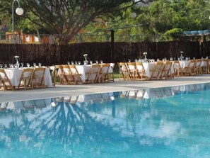 A poolside dining area with tables set for a meal, surrounded by a wooden fence and trees. - Elifaz Kibbutz Desert Hospitality (Hevel Eilot)