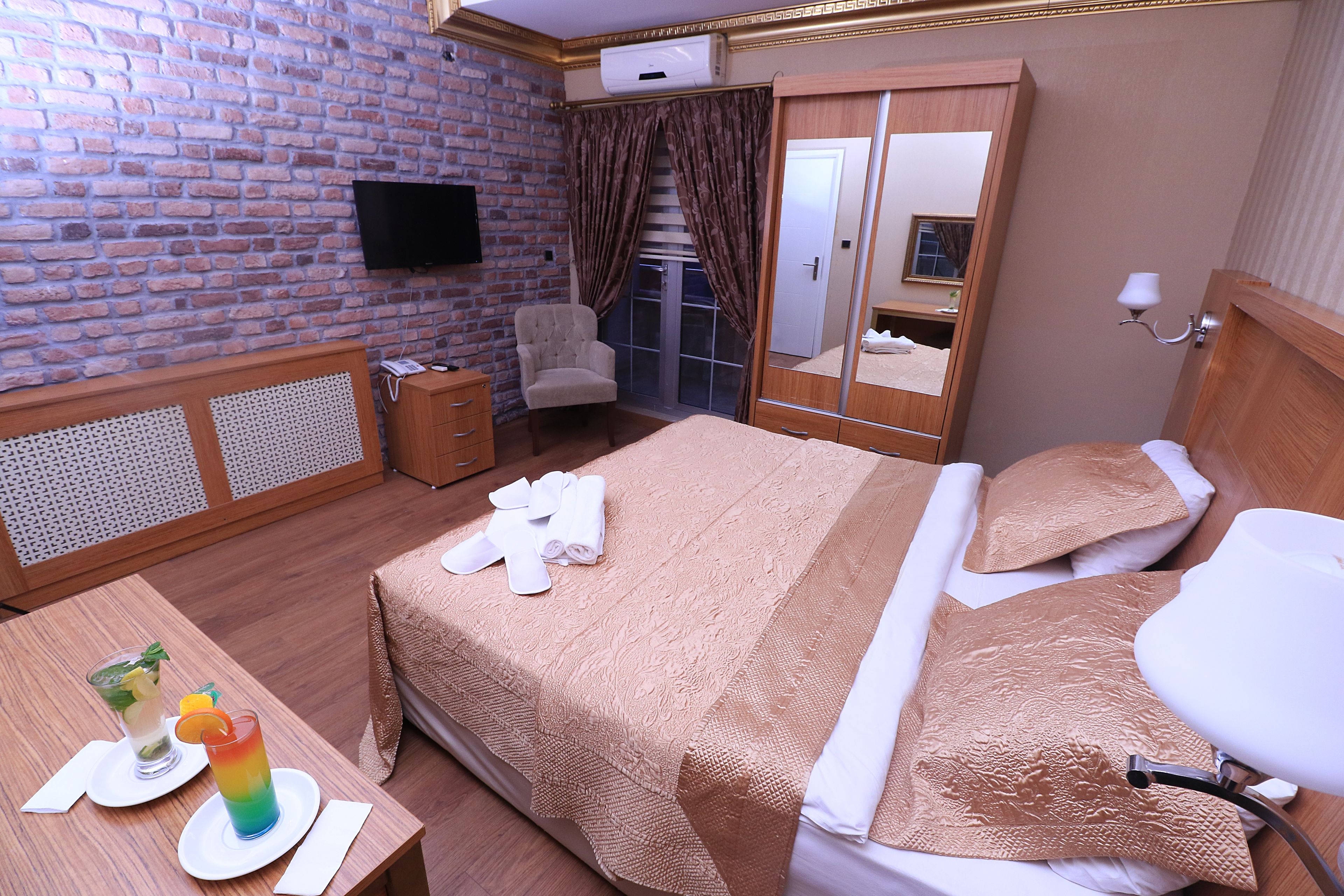 Habitación de hotel con pared de ladrillo, televisor de pantalla plana, cama con cubrecama color beige, mesita de noche de madera con un vaso de agua y una tajada de fruta, y un espejo con marco de madera.